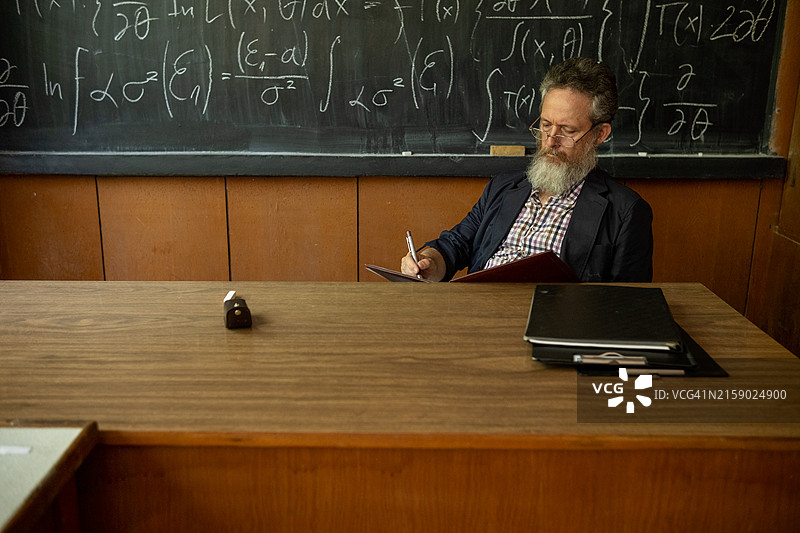 一位留着长白胡子、灰色大头发、戴着眼镜的老师，穿着格子衬衫和运动夹克，站在写有复杂公式的黑板前。图片素材