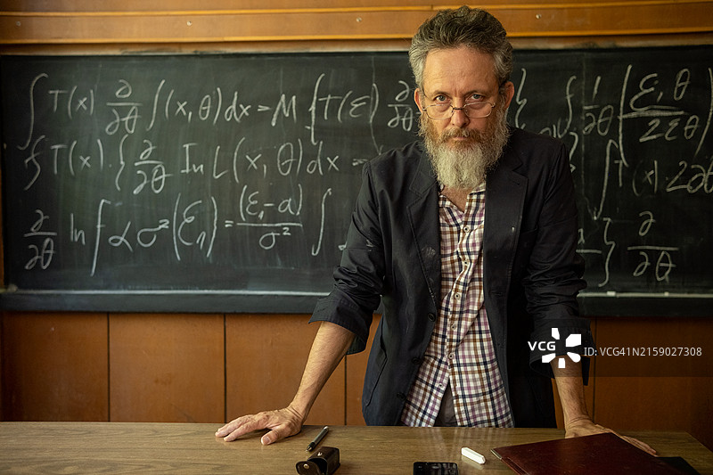 一位酷老师的肖像，他有着长长的白胡子、大灰发和眼镜，穿着格子衬衫和运动夹克，站在写有复杂公式的黑板前。图片素材