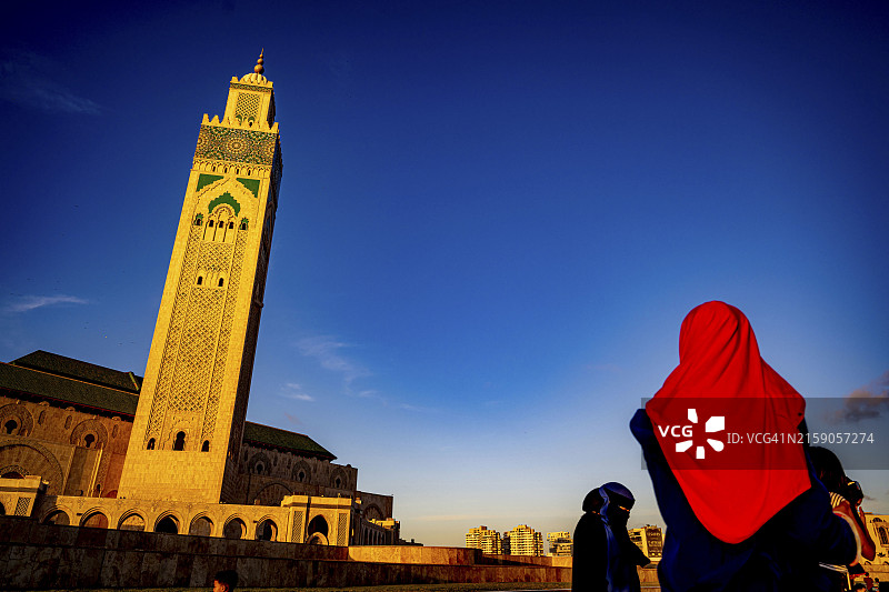 夕阳下，戴着鲜艳红色头巾的人凝视着雄伟的清真寺塔，背景是深蓝色天空图片素材