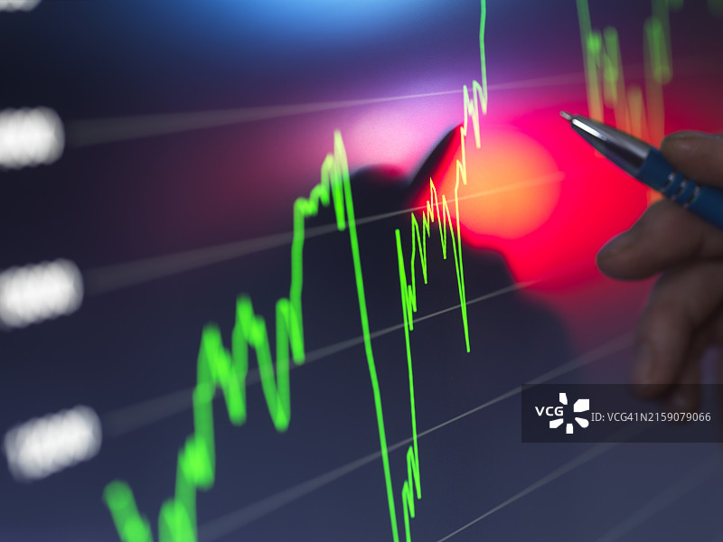 手握笔指向数字屏幕上明亮的股市图表，绿色和红色线波动，指示金融数据图片素材