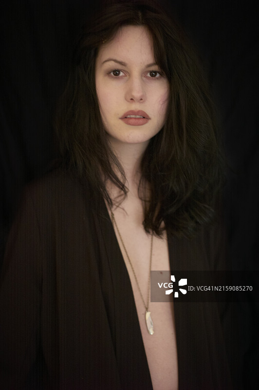 一位头发蓬乱、表情平静的年轻女子身穿深色开衫，佩戴白色吊坠项链，黑色背景突显她的存在图片素材