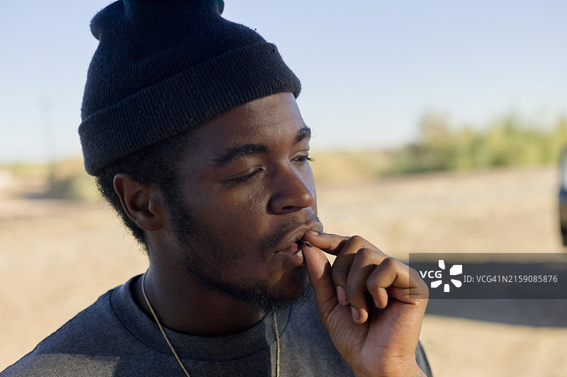 阳光下，戴着无檐小便帽的年轻男子若有所思地咬着指甲图片素材