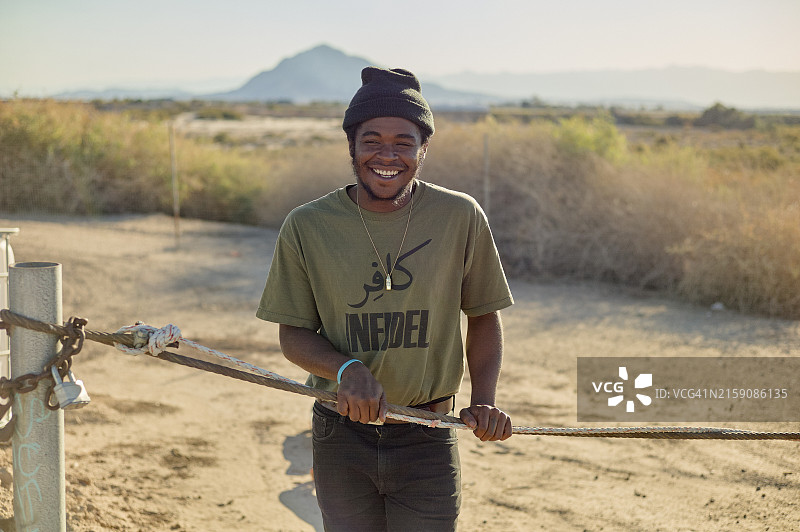 面带微笑的男人戴着无檐便帽和绿色T恤站在金属门后，背景是阳光明媚的山景图片素材