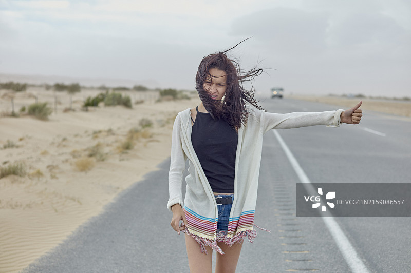 年轻女子站在沙漠公路旁，长发被风吹乱，竖起拇指搭便车图片素材
