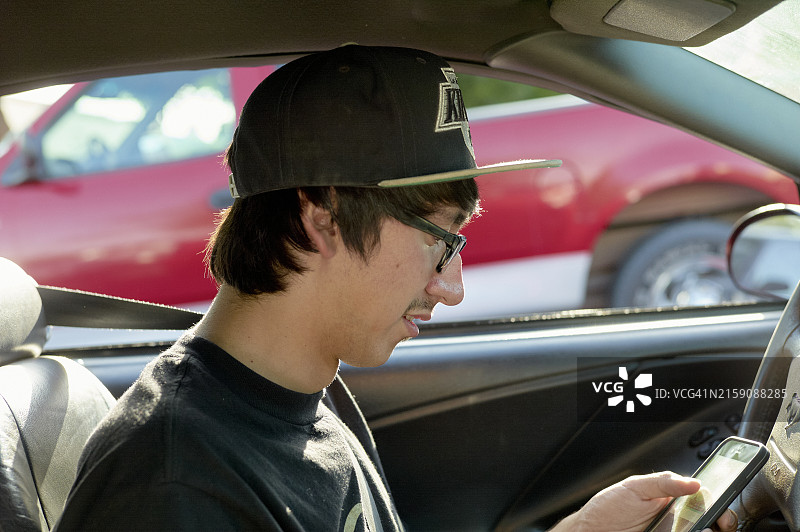 戴眼镜棒球帽的年轻男人坐在驾驶座上，低头看手机微笑图片素材