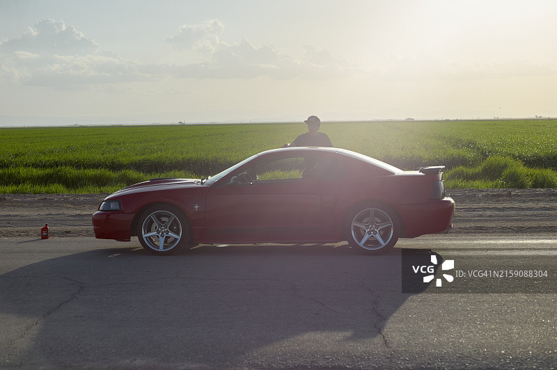 一个人站在停在路边的红色跑车的车顶天窗里，背景是茂盛的绿色田野和晴朗的天空图片素材