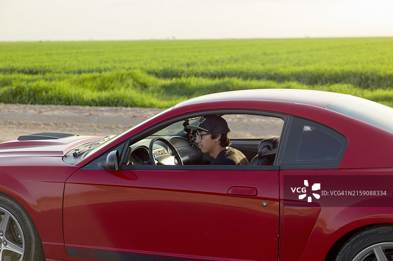 戴着太阳镜和帽子的人驾驶红色跑车穿过绿色田野的乡村地区图片素材