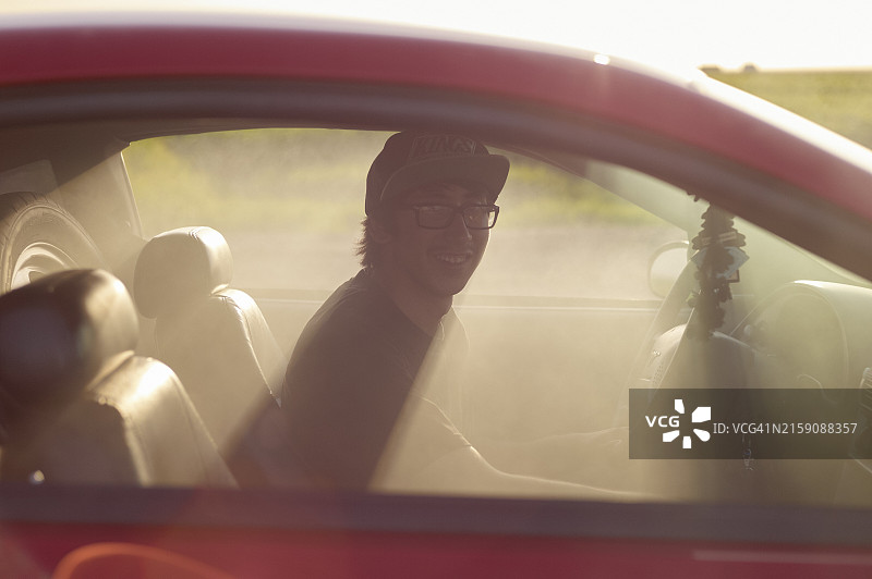 戴棒球帽和眼镜的男人坐在阳光明媚的汽车驾驶座上微笑图片素材