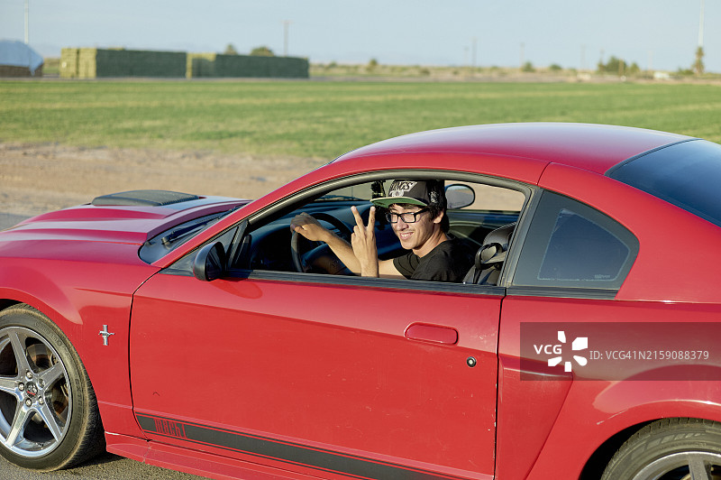 在一个阳光明媚的户外区域，男人从红色跑车的驾驶座上伸出大拇指，面带微笑图片素材