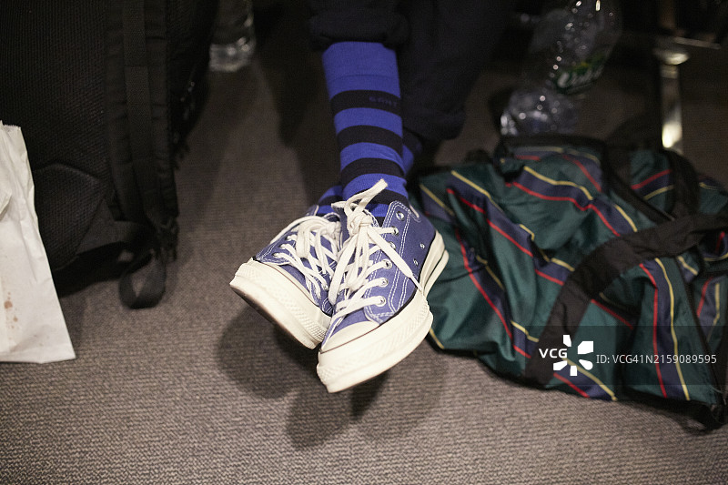 穿着蓝色袜子和白色蓝色鞋带运动鞋的人的脚部特写，旁边是地毯上的绿色条纹旅行包图片素材