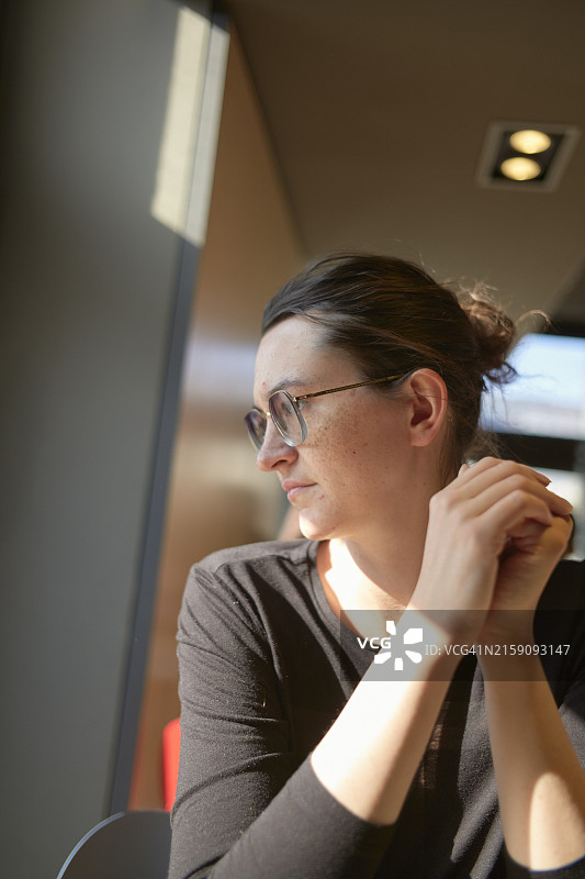 戴眼镜的女子在室内沉思，手托下巴，沐浴在自然光中图片素材