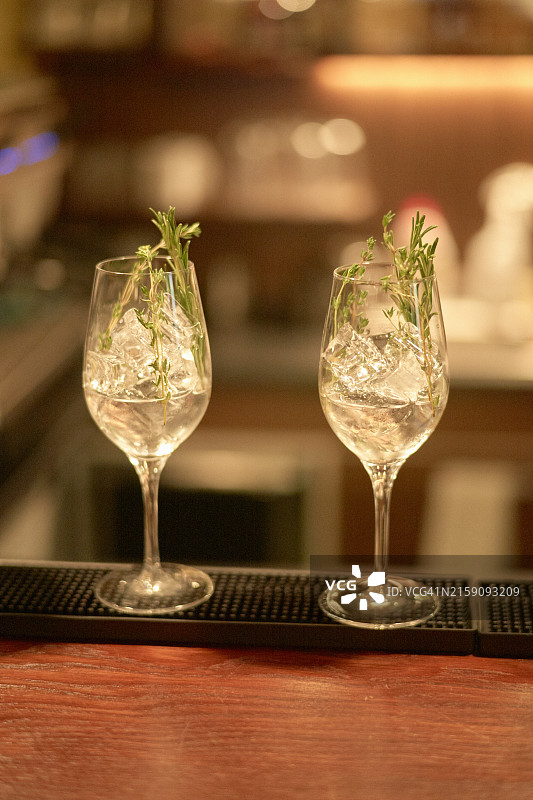 两个优雅的玻璃杯中盛满透明液体，并用小枝香草装饰，摆放在吧台上，背景柔和模糊图片素材