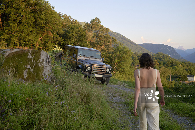 在黄昏时分走向停在乡村公路旁的越野车的人，背景是茂盛的绿色植物和山脉图片素材