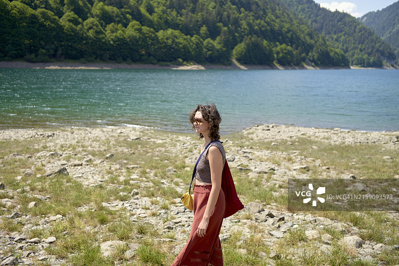 红裙女人走在风景优美的湖边岩石岸边，四周环绕着蓝天下森林覆盖的丘陵图片素材