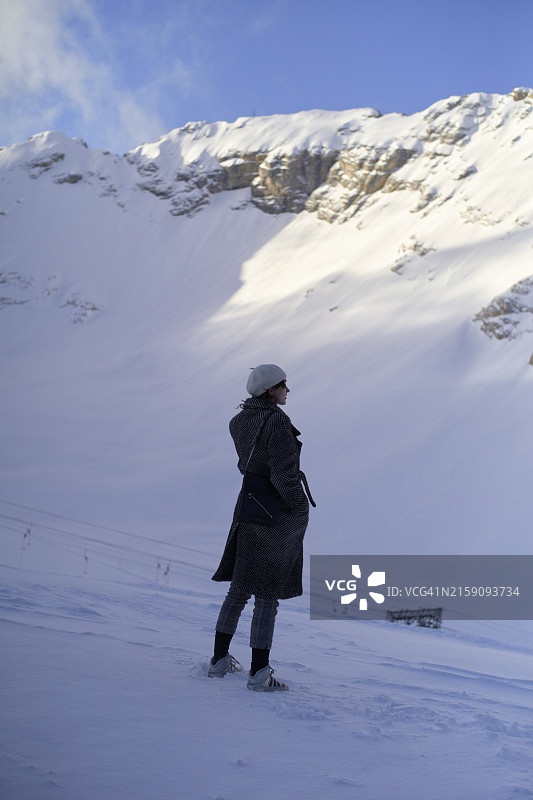 在晴朗的蓝天下，一个人站在白雪皑皑的风景中，凝视着高耸的阳光照耀的山脉图片素材