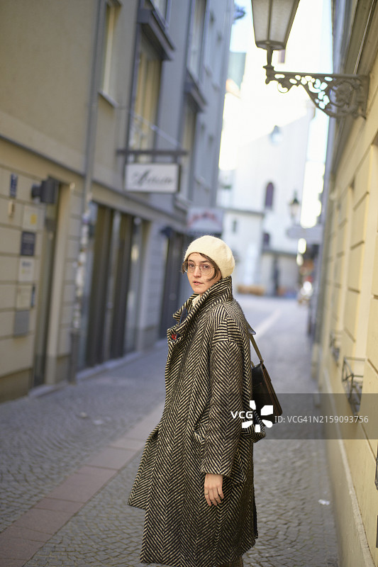 身穿人字纹外套和白色贝雷帽的人站在狭窄的小巷里，侧身回望图片素材