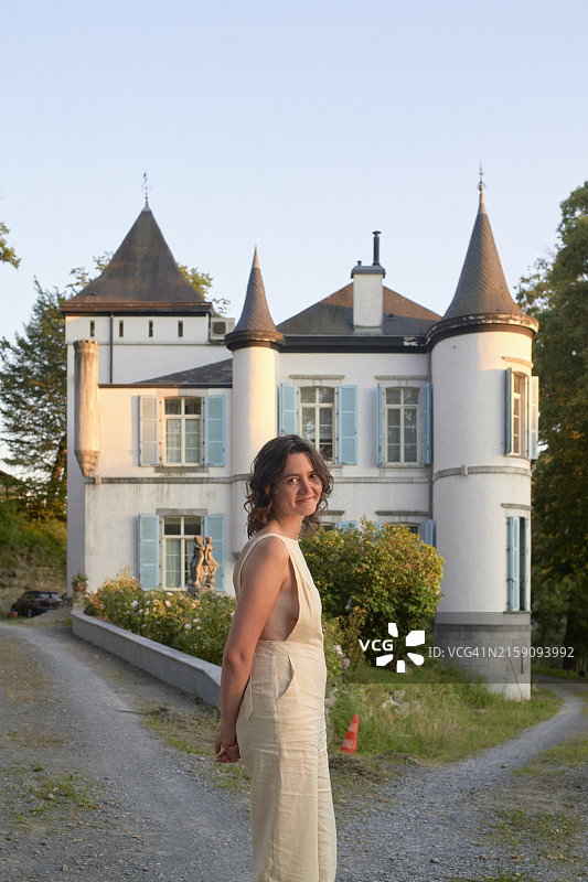 一位穿着时尚奶油色套装的微笑女人站在优雅的白色城堡前，城堡有双塔和茂盛的绿色植物图片素材