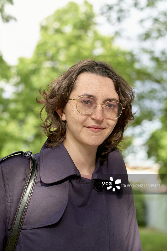 戴着眼镜，穿着紫色上衣，留着棕色齐肩发的年轻微笑女子，背景是树图片素材