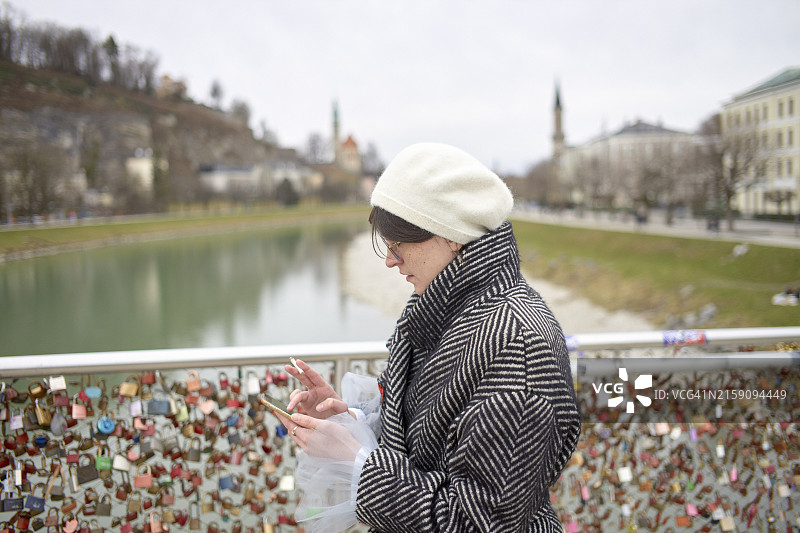 站在桥上俯瞰河流，桥的栏杆上挂满了爱情锁，同时查看智能手机的人图片素材