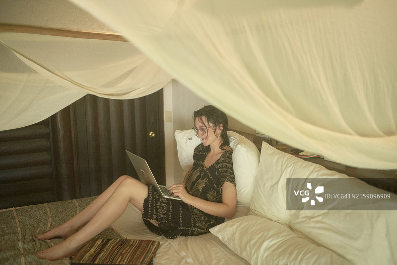 女人坐在铺着白色床单的床上，正在使用她的笔记本电脑。她在一张柔软、透明的蚊帐下，戴着眼镜，穿着休闲服装，在一个舒适、光线昏暗的房间里姿势放松图片素材