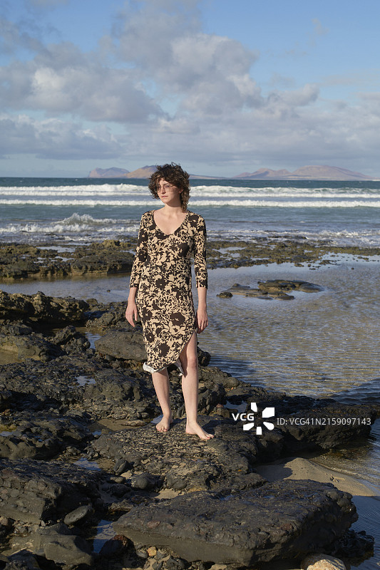 年轻女子站在岩石海滩上，穿着碎花裙，背景是海洋和岛屿图片素材