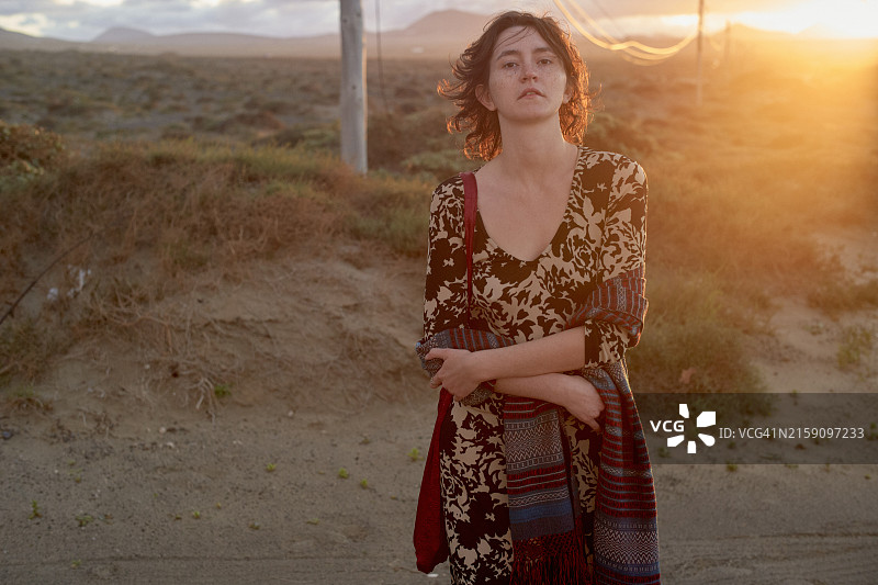 日落时分，站在户外的沉思的女子，穿着碎花连衣裙，搭配开衫，手臂上搭着花纹毯子，背景是沙漠景观图片素材