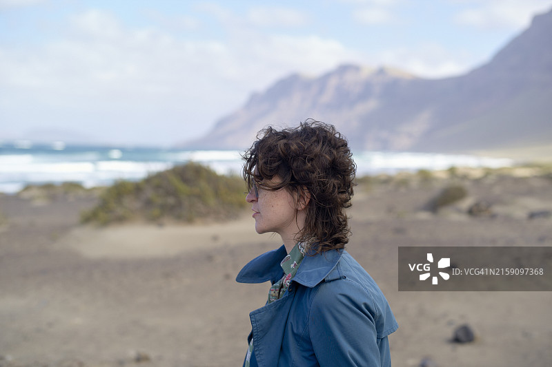 站在海滩上眺望远方山脉的卷发年轻女子图片素材
