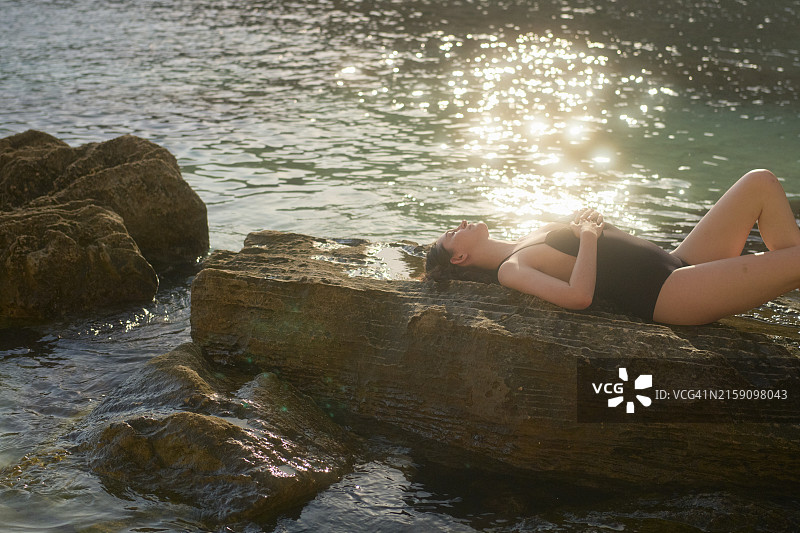 年轻女子在海边岩石上放松身心，享受阳光图片素材