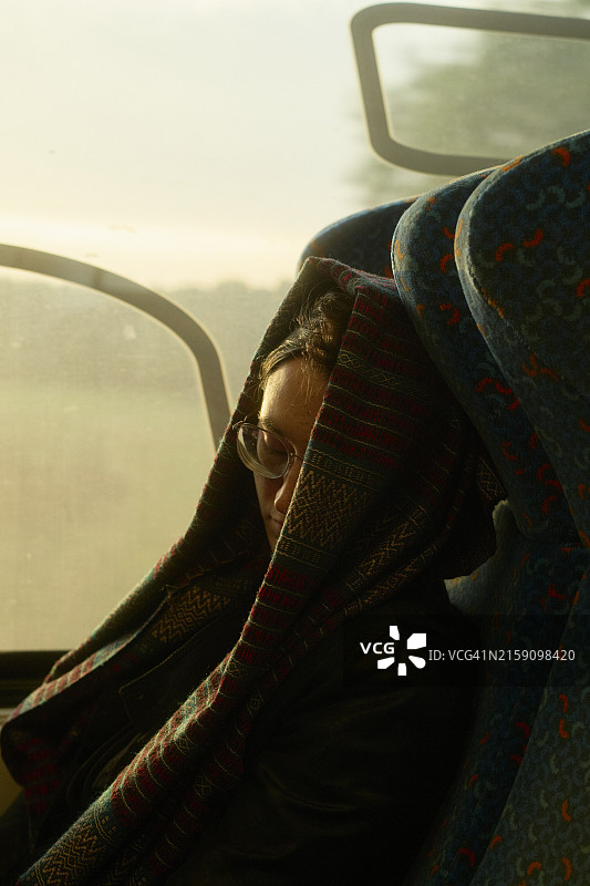 年轻女子坐在公共汽车上，脸部被彩色围巾部分遮挡，阳光透过窗户图片素材