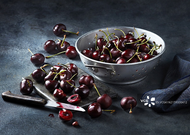 一碗新鲜的樱桃，深色纹理表面，周围散落着一些樱桃，其中一个被咬了一口图片素材
