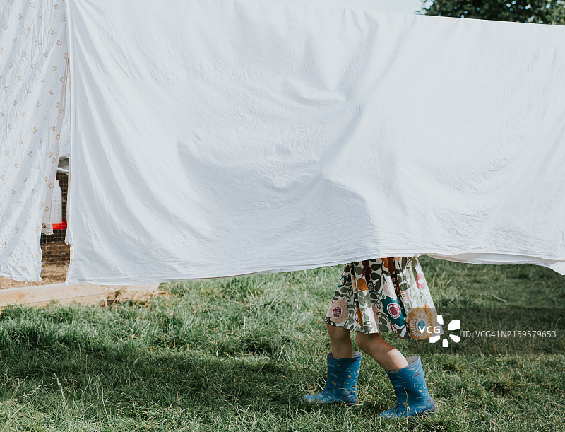花园里，孩子躲在晾衣绳上悬挂的白色床单后，只露出双脚图片素材
