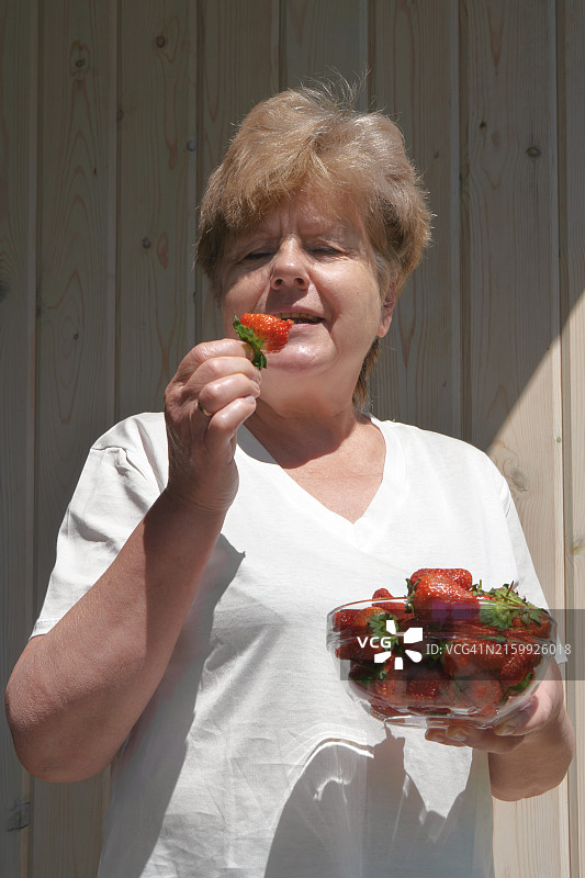 阳光明媚的日子里，穿着白色T恤，手捧一碗浆果，吃新鲜草莓的快乐女人肖像图片素材