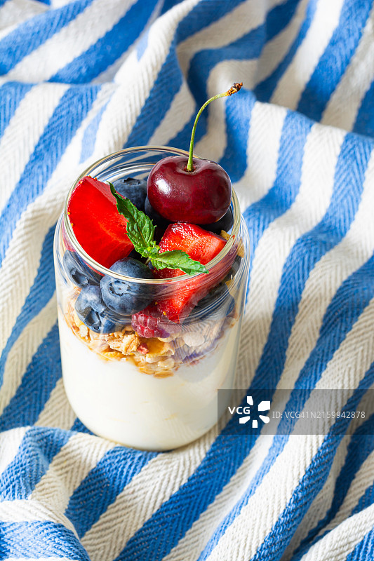 白色和蓝色背景的玻璃罐中盛有格兰诺拉麦片和新鲜水果的酸奶，健康早餐零食，外卖概念图片素材