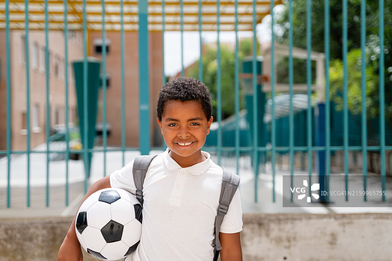 微笑的小学生在学校拿着足球，背着书包图片素材