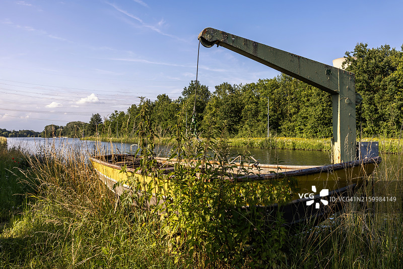 悬挂在河岸起重机上的旧的、杂草丛生的划艇图片素材