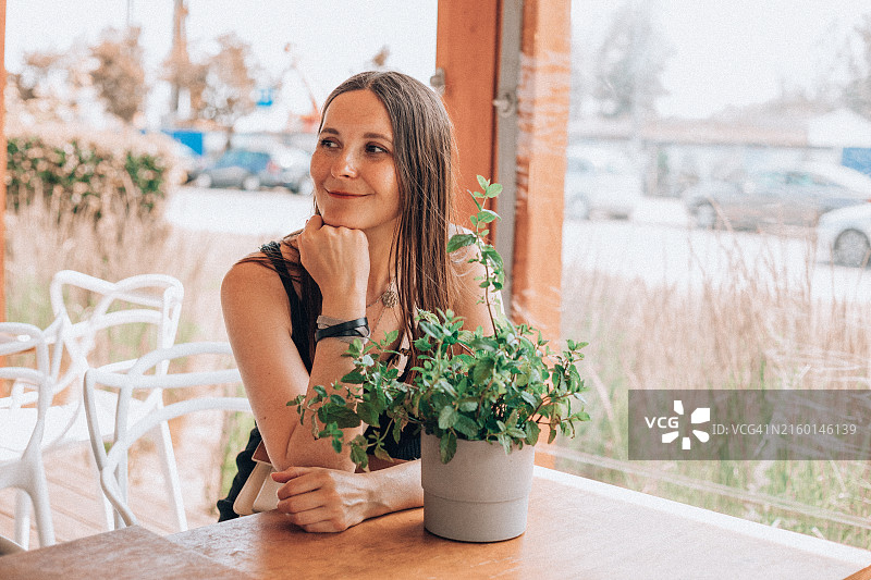 坐在桌旁，手托下巴，面带微笑地望着远方，在咖啡馆阳台上享受宁静的迷人女人图片素材