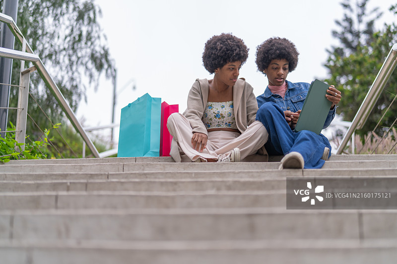 姐妹坐在楼梯上休息，使用平板电脑，旁边放着礼品袋图片素材