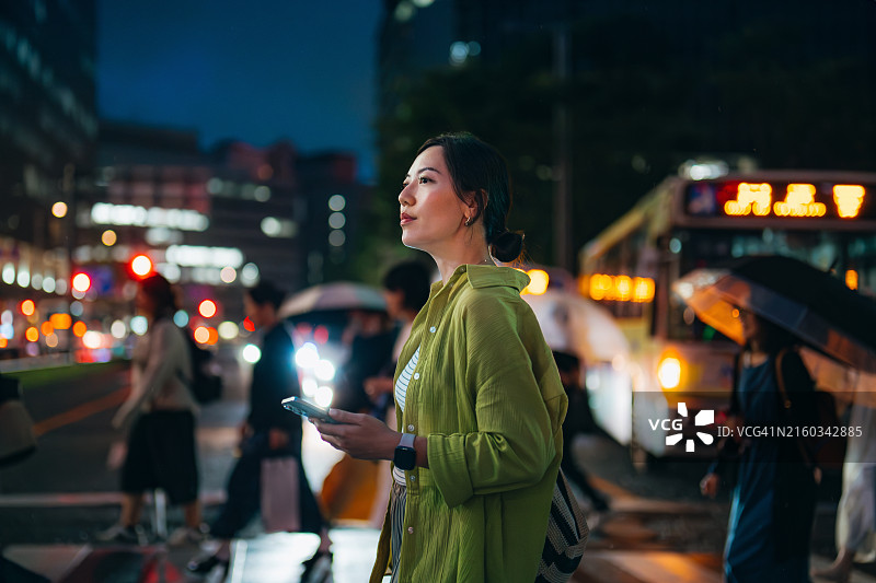夜晚在繁忙的城市街道用手机过马路的年轻亚洲女性图片素材