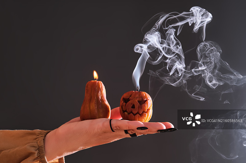 做黑色美甲、戴戒指的女人手拿两支点燃的万圣节南瓜造型蜡烛。照片展示了蜡烛熄灭后的大气白色烟雾。图片素材