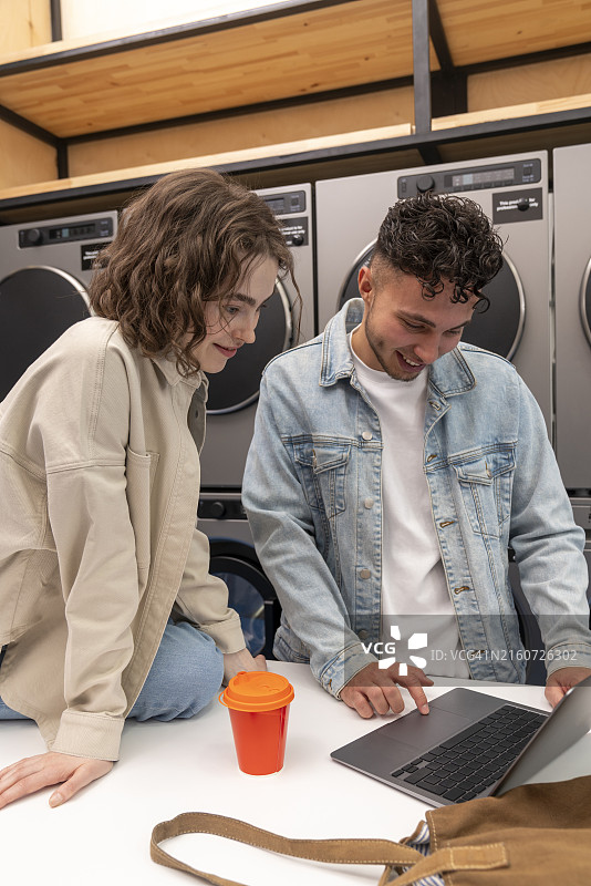 微笑情侣在自助洗衣店用笔记本电脑图片素材