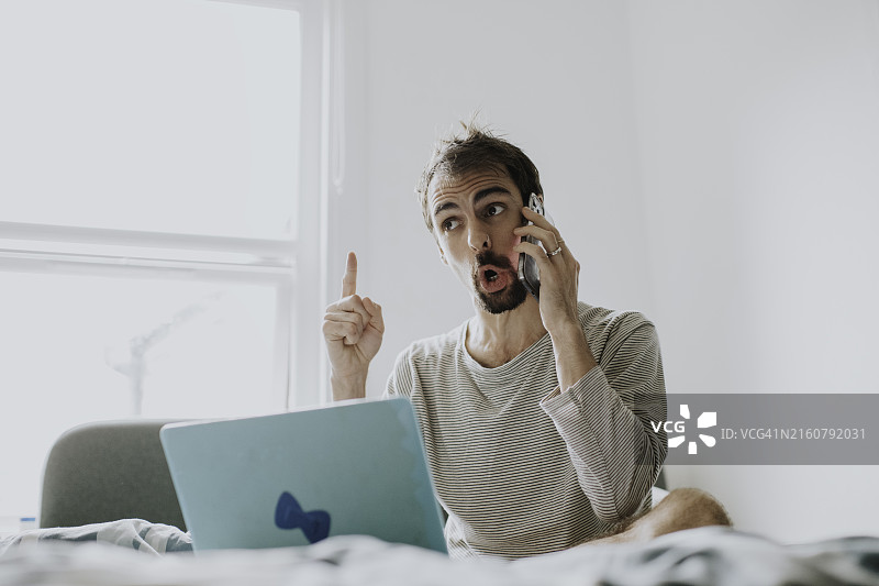 一个沮丧的男人坐在床上，拿着笔记本电脑，一边打电话一边表达不满图片素材