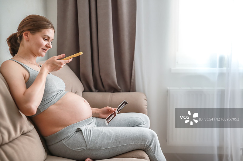 怀孕的女人在家中沙发上进行视频聊天，同时在智能手机上展示婴儿的超声波照片图片素材