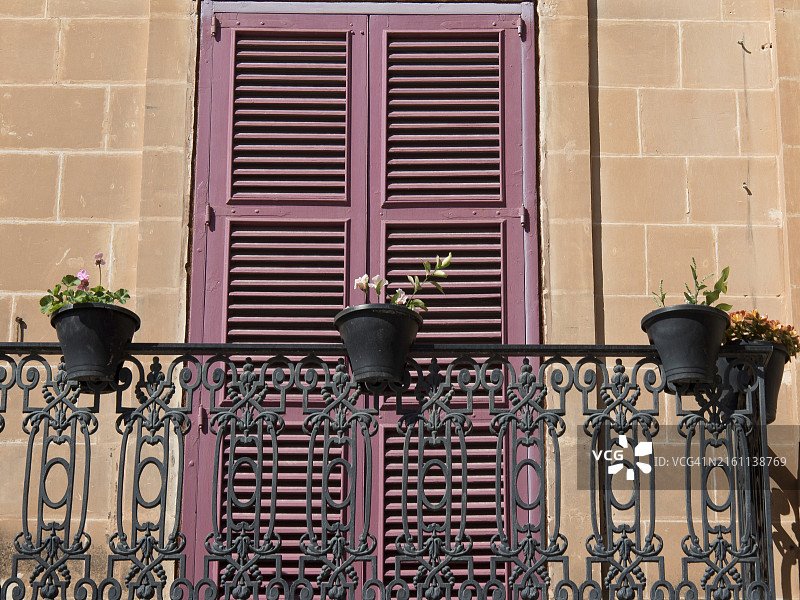 带有锻铁栏杆和粉色百叶窗的阳台，饰有多个花盆，位于马耳他姆迪纳地中海的砂岩外墙前图片素材