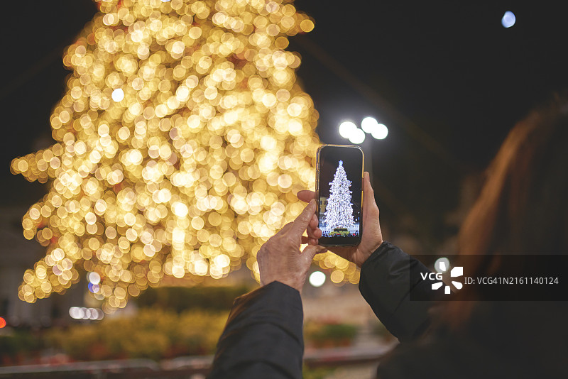 女子在户外用手机拍摄灯光璀璨的圣诞树的背影图片素材