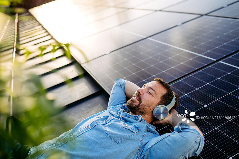 躺在装满太阳能电池板的屋顶上，戴着无线耳机听音乐的男人。可持续生活方式、光伏和可再生能源概念图片素材