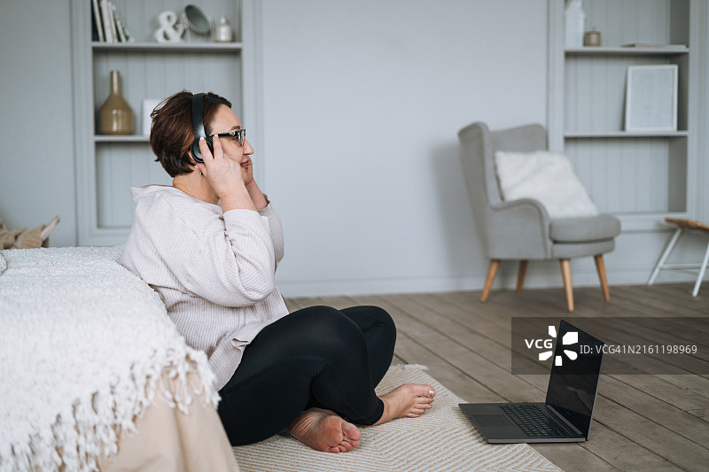 在卧室里戴着耳机用笔记本电脑听音乐的加大码中年妇女图片素材