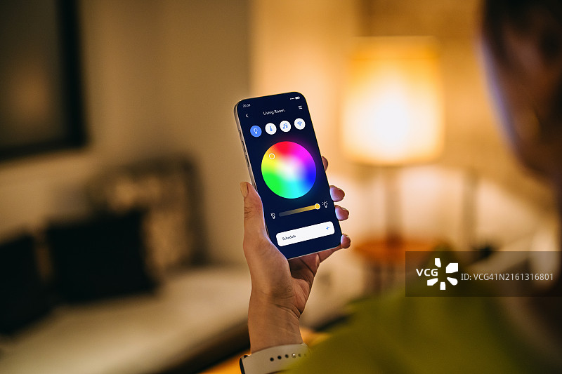 肩上视角：女人通过智能手机上的移动应用程序设置智能家居系统，调整智能家居的照明和家用电器。家庭自动化。生活方式和技术。智能家居技术概念图片素材