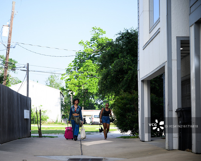 Z世代女性朋友拉着行李箱走在绿树成荫的郊区道路上，到达度假出租屋图片素材