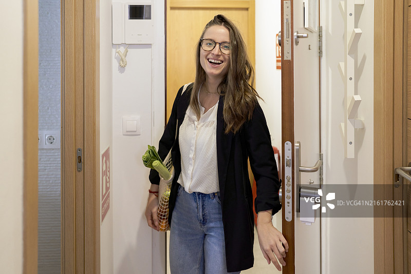 一位戴眼镜的美丽女人打开公寓的门，手里拿着一袋蔬菜，面带微笑地看着镜头，正面视角图片素材