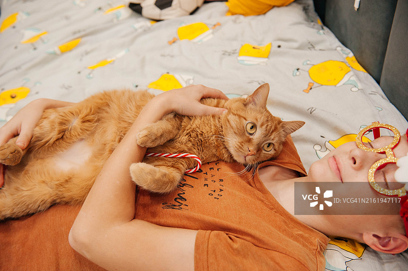 戴着新年眼镜、拿着新年装饰品的男孩抱着一只姜黄色的猫。图片素材